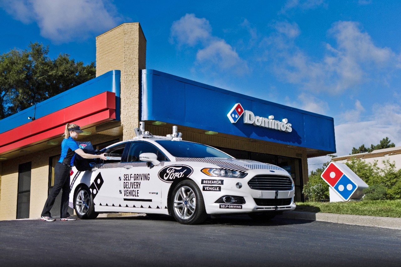 A white Ford Fusion with visible self-driving hardware is shown in front of a Domino's Pizza restaurant. A Domino's worker is loading a pizza into the vehicle. 