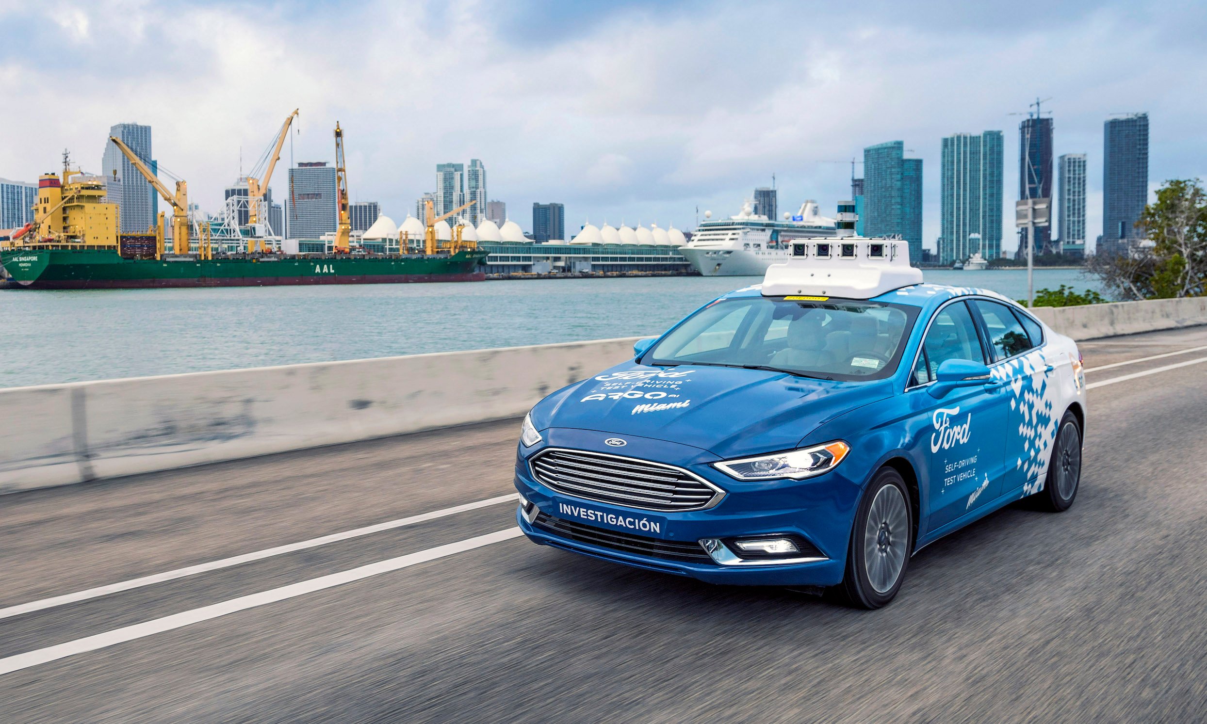 A blue and white Ford Fusion sedan with visible self-driving hardware on a street in Miami, Florida.