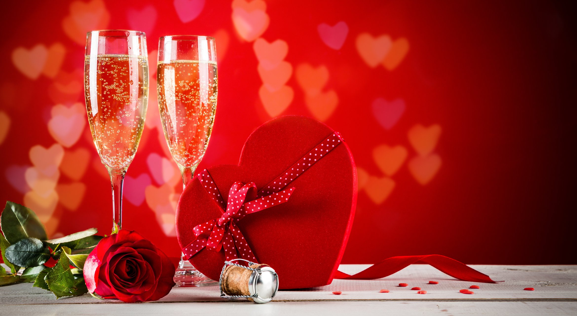A table has champagne glasses, a box of heart-shaped chocolates, and a rose.
