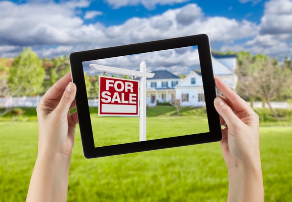 Female hands holding computer tablet with "home for sale" sign on screen, house behind.