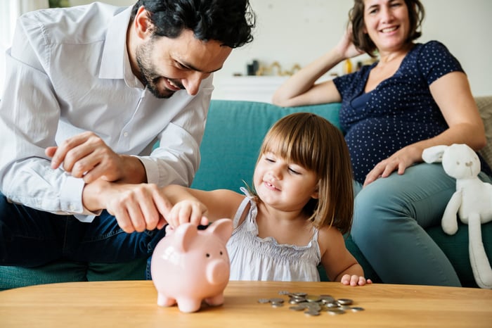 Young girl inserts coin into piggy bank while her father and pregnant mother look on.