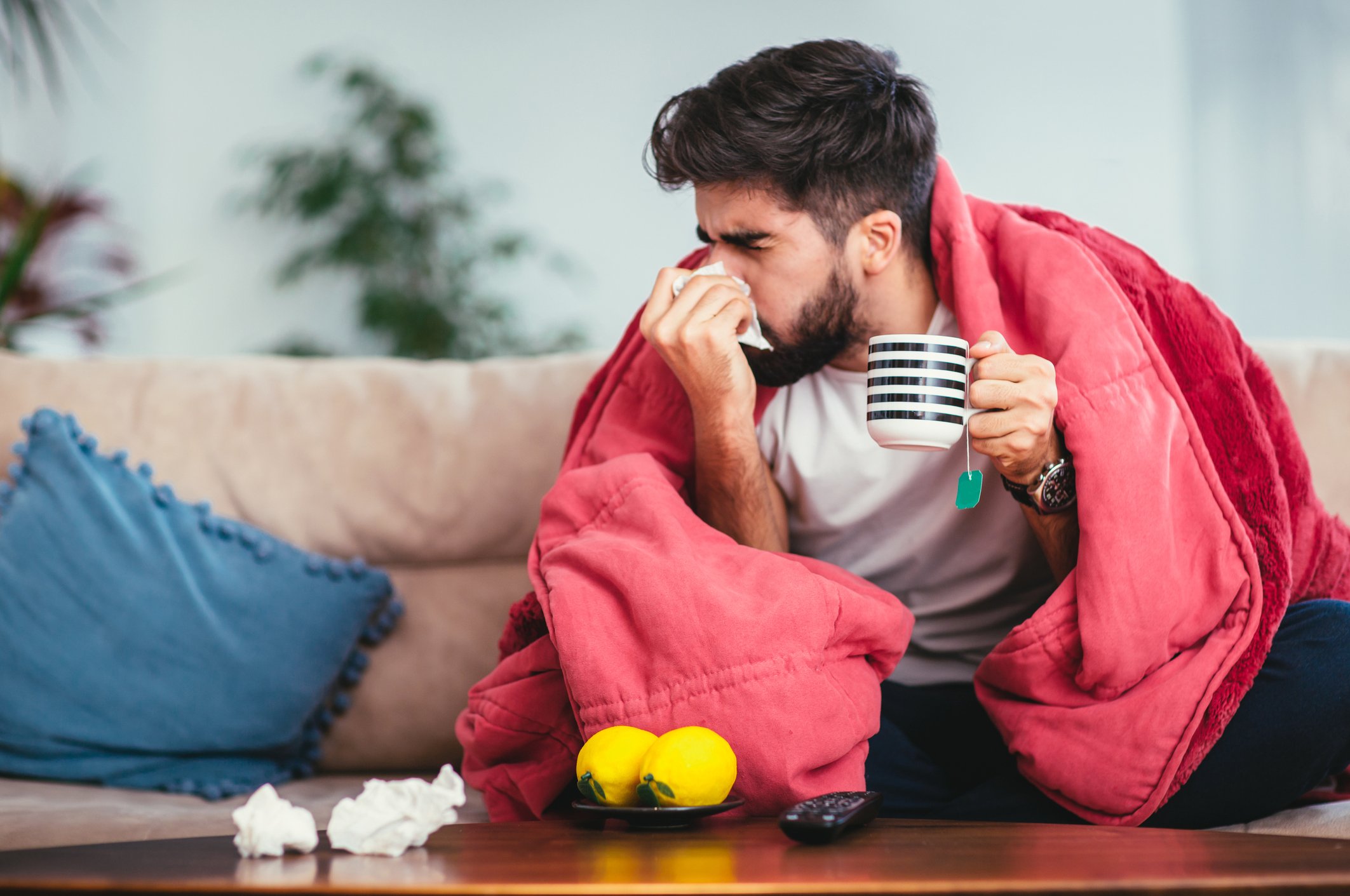 A man wrapped in a blanket and holding a mug of tea blows his nose into a tissue.