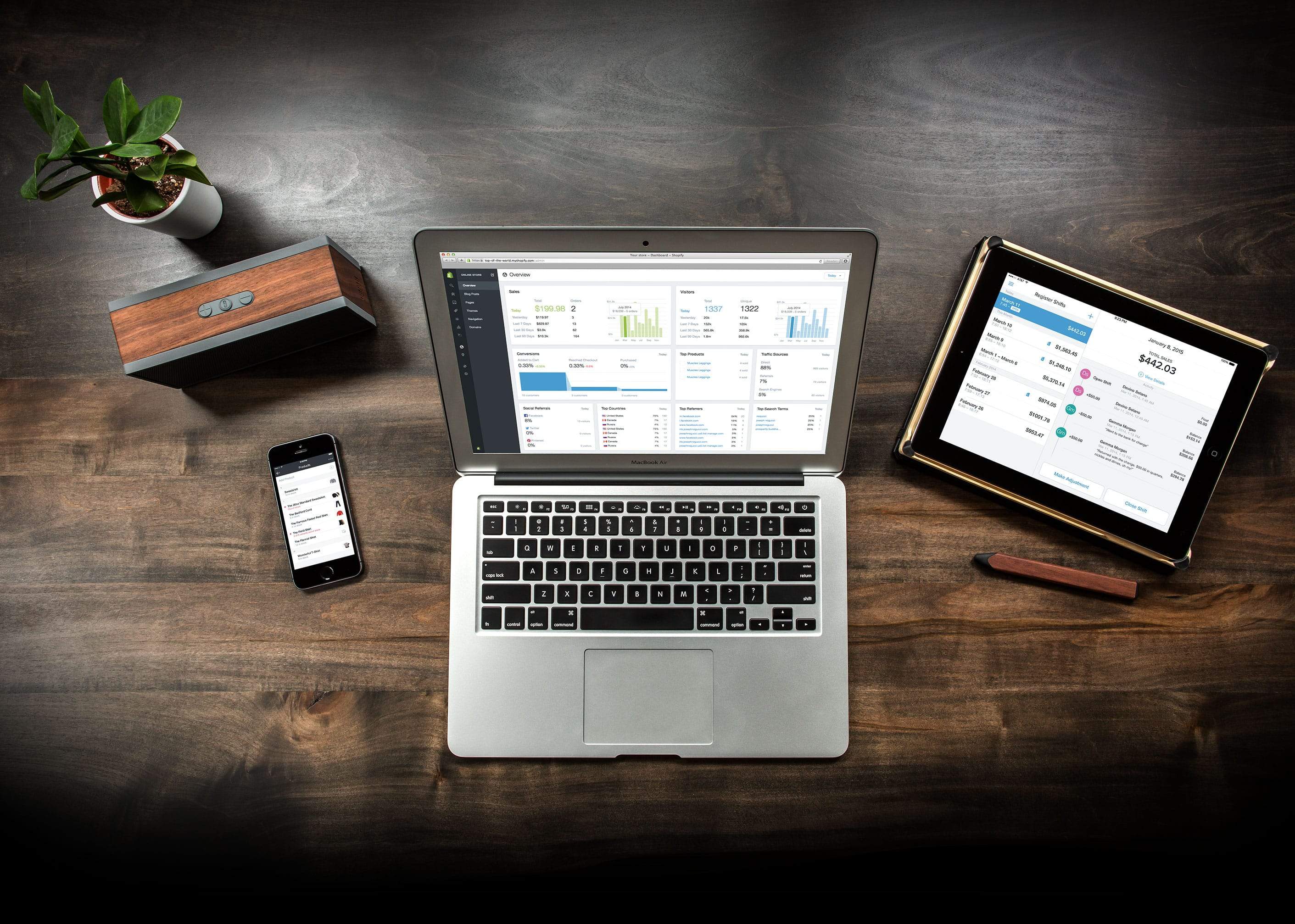 A wooden desk with a laptop, notebook, and smartphone -- all displaying the Shopify app.