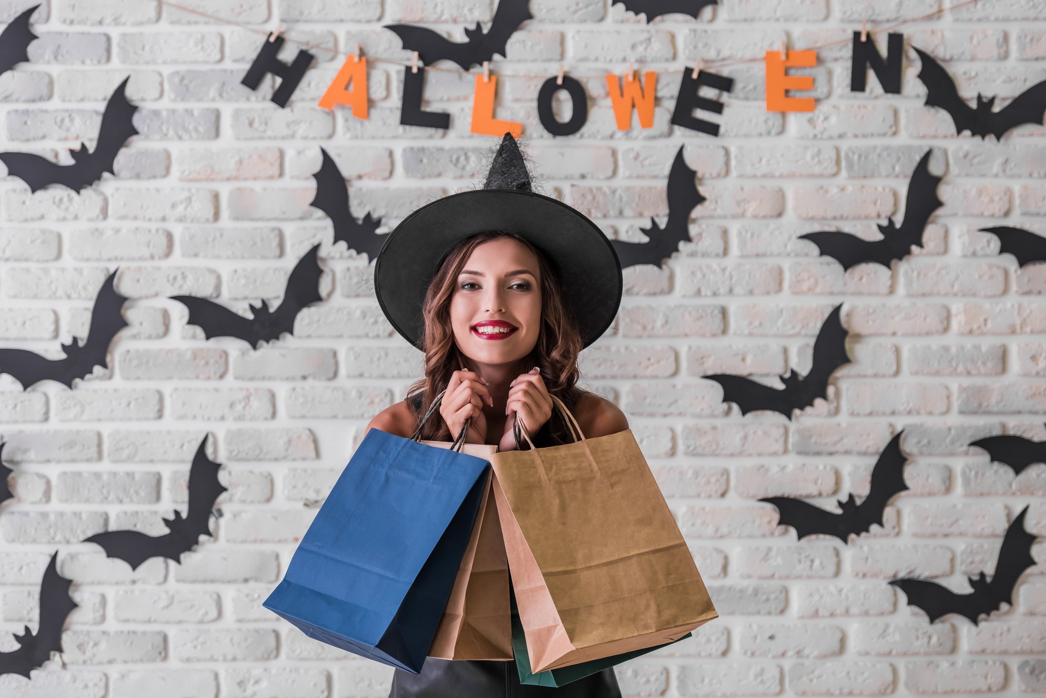 A woman wearing a witch hat holds up shopping bags.