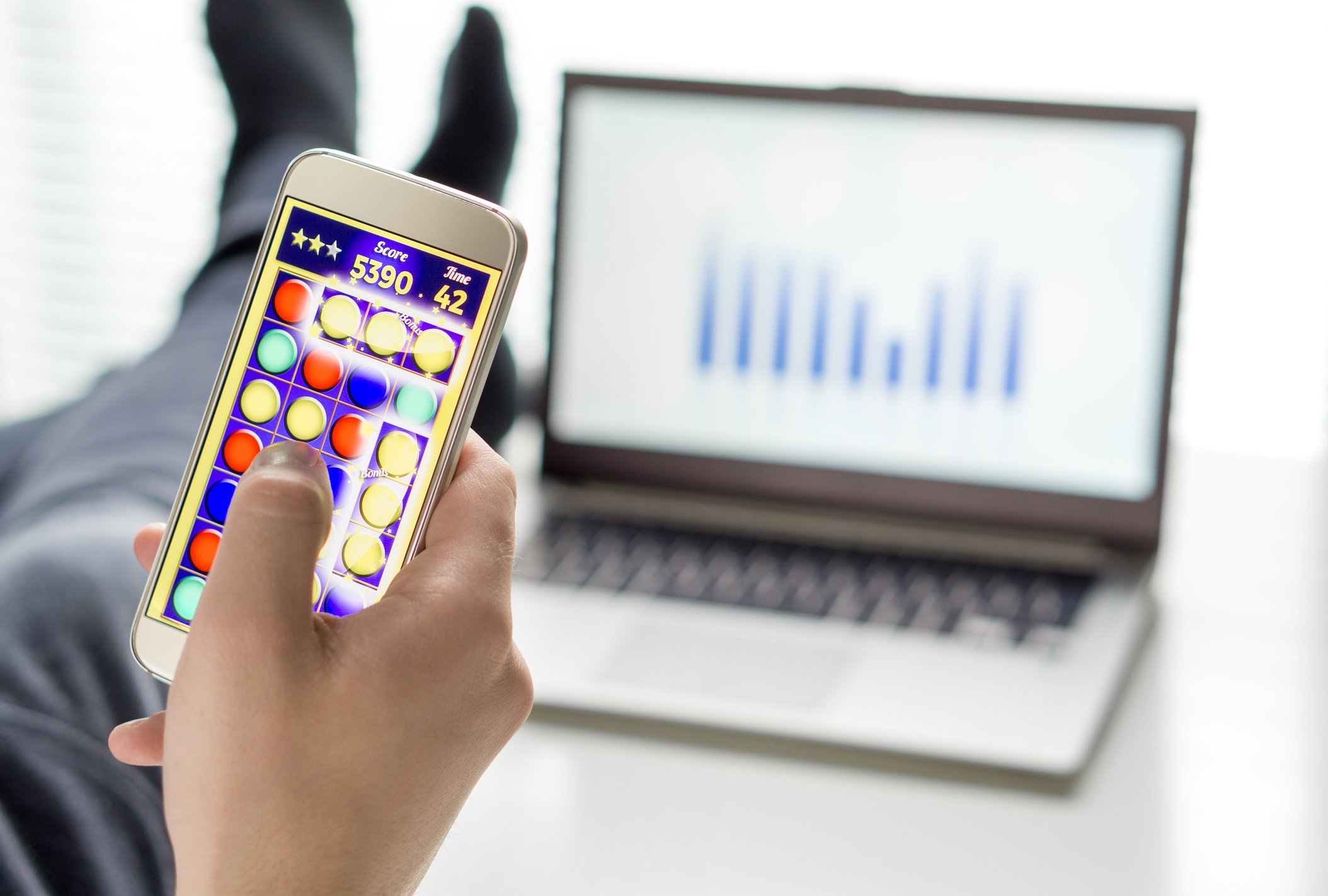 Man playing a mobile game with a work laptop in the background.