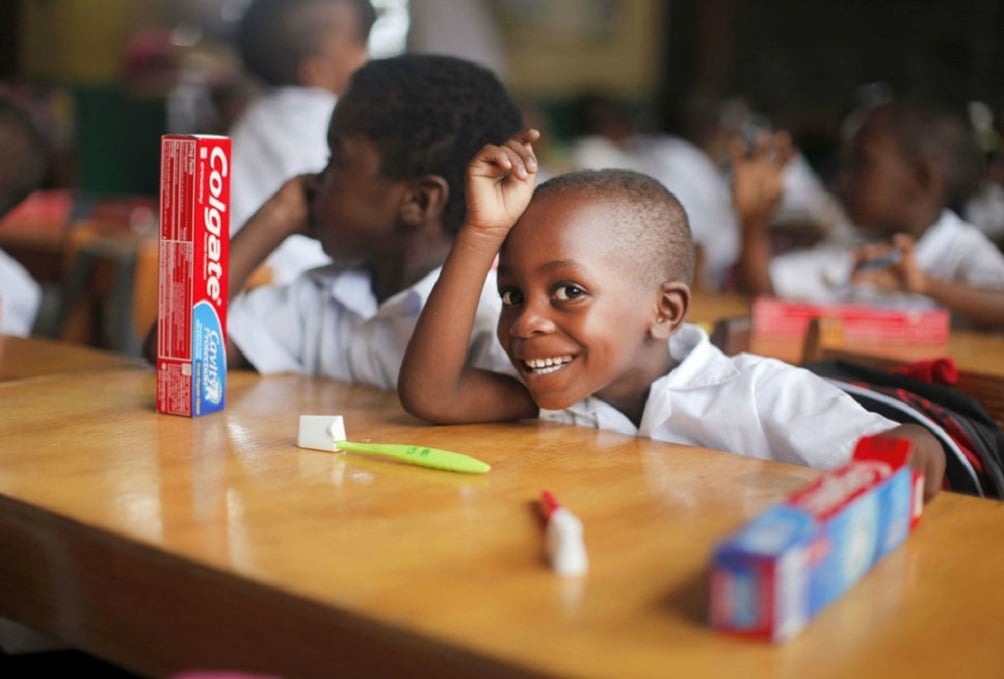 Child in a room with other children smiling, with a toothbrush and Colgate toothpaste on a table in front of the child.
