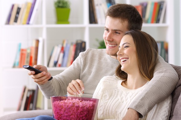 A smiling young couple share a bowl of popcorn, TV remote in hand.