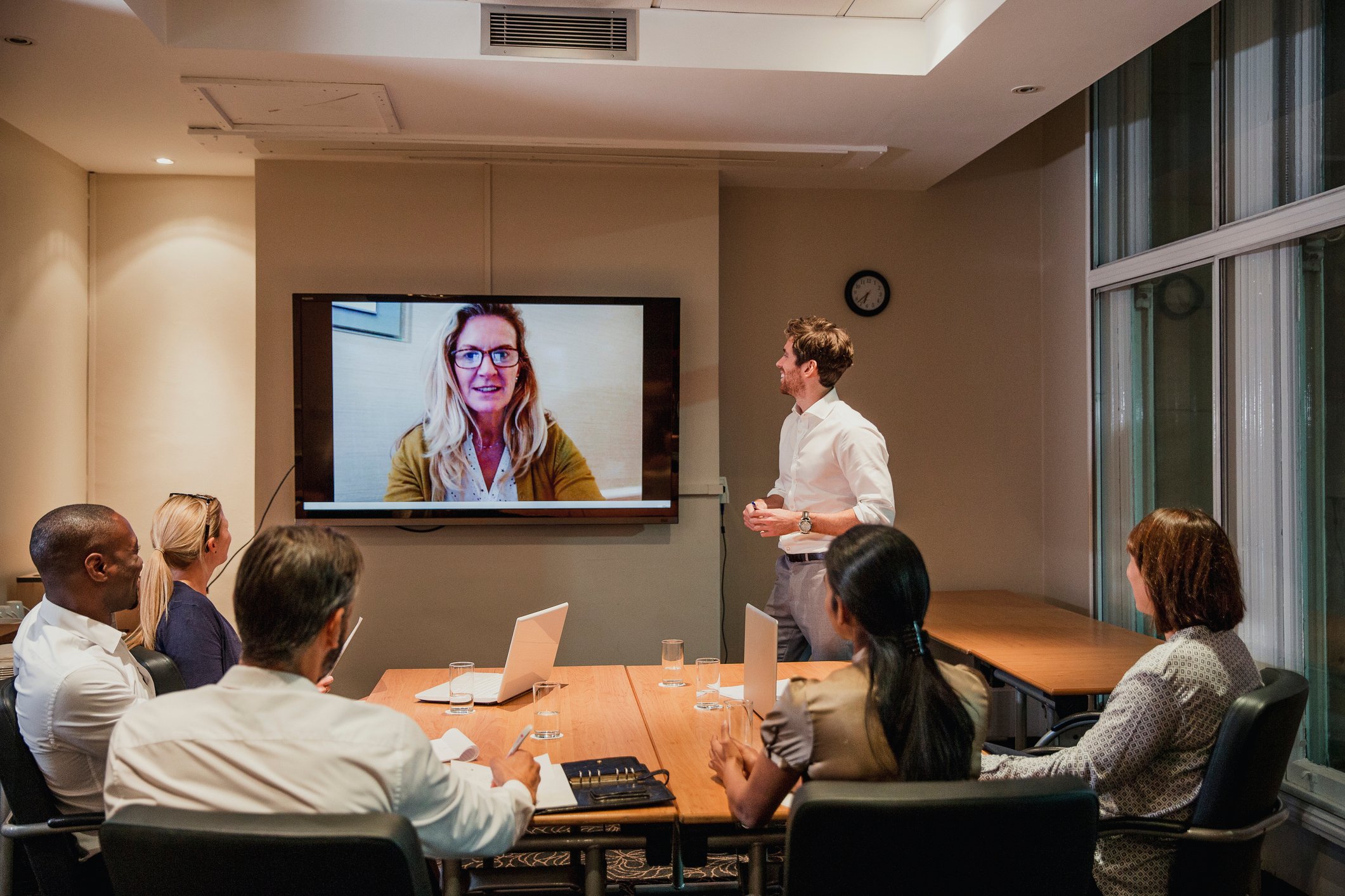 Professionally dressed adults in a conference room looking up at a woman videconferencing in on a large screen.