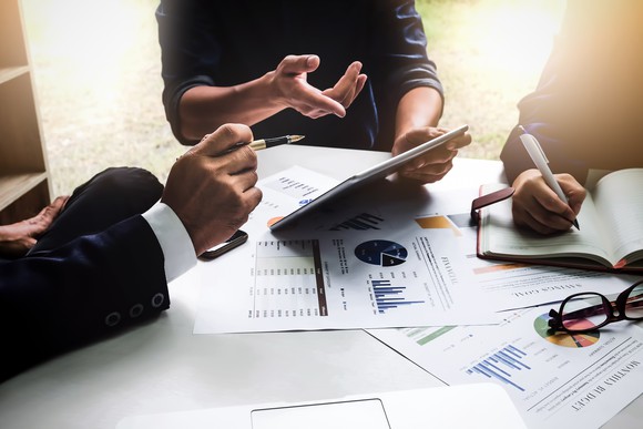 Business people sitting around a table, analyzing charts, while one person points at something on a tablet computer.