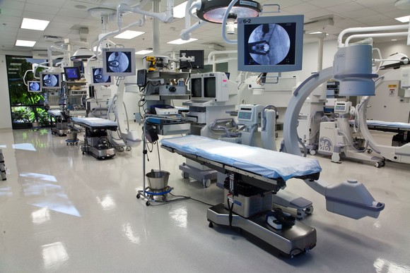 Several spinal surgical tables with imaging equipment and various monitors nearby, in a room with white tile floors and bright fluorescent lighting.