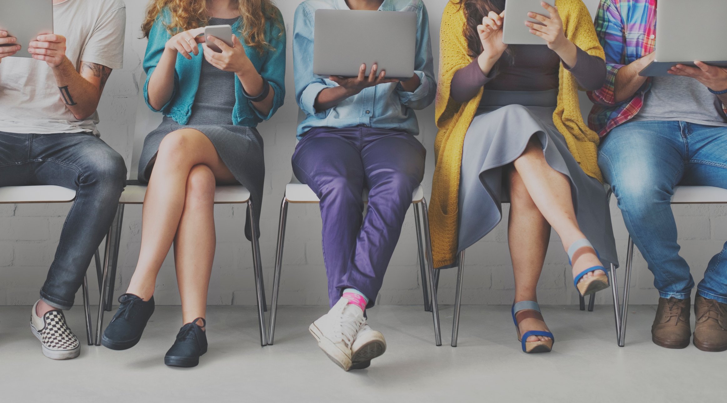 Young people using mobile devices while sitting on chairs lined up.