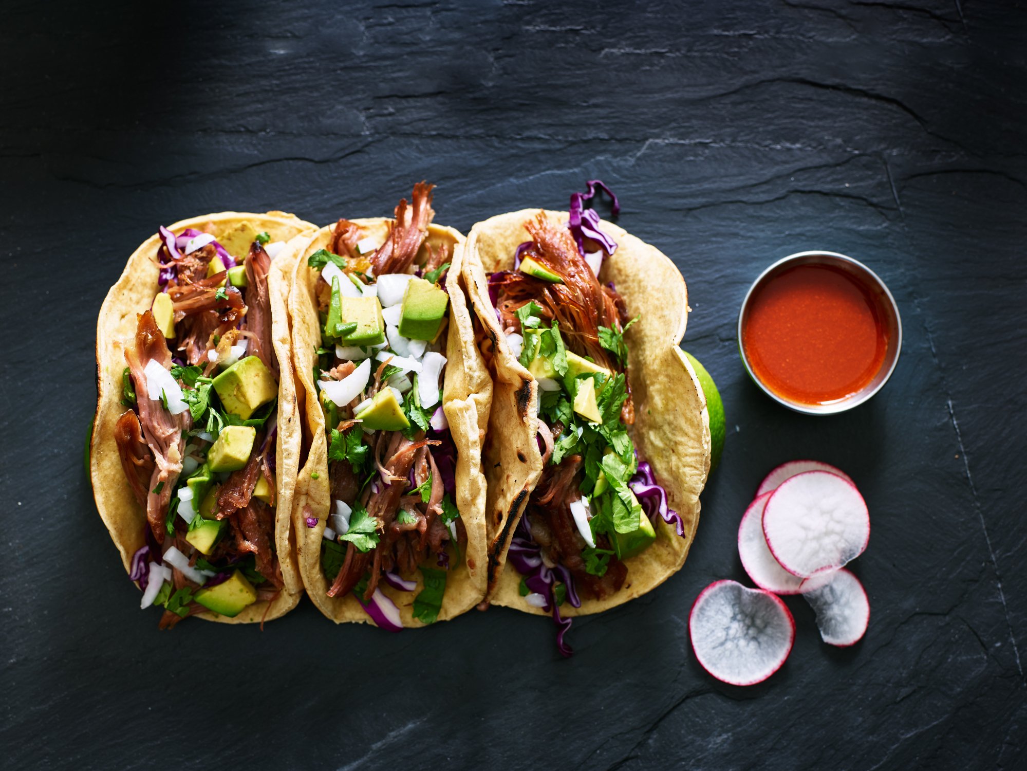 Three tacos on a table next to sauce and radishes.