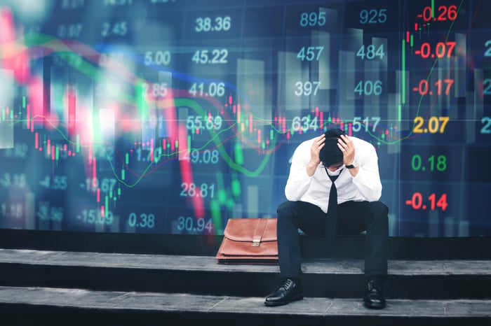 With his head in his hands, a businessman sits in front of a digital financial chart featuring a large red line and various numbers.