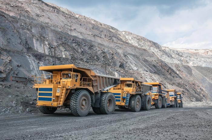 Several trucks drive in a line up a ramp in a mining area.