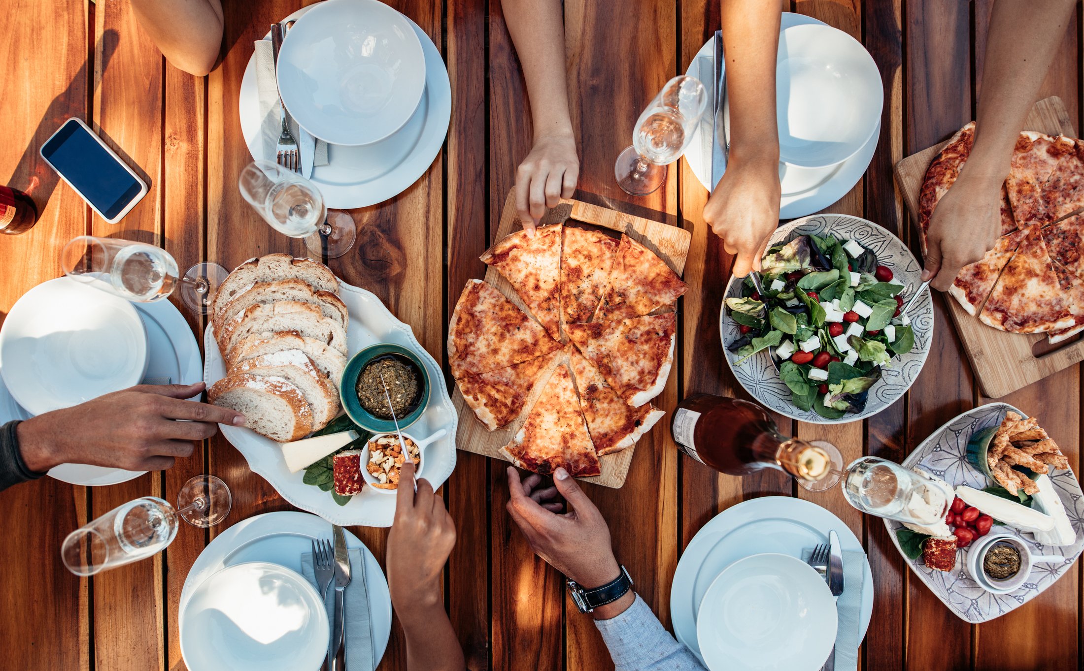 A table with five place settings and six hands reaching out to grab pizza, salad, bread, and other food at the center of the table.