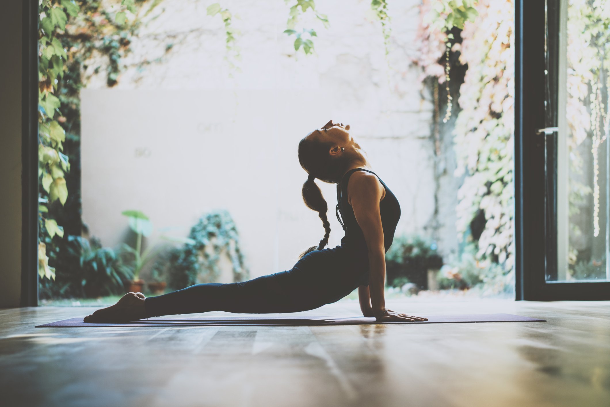 A woman holding a yoga pose in front of doors opened to courtyard with various plants.