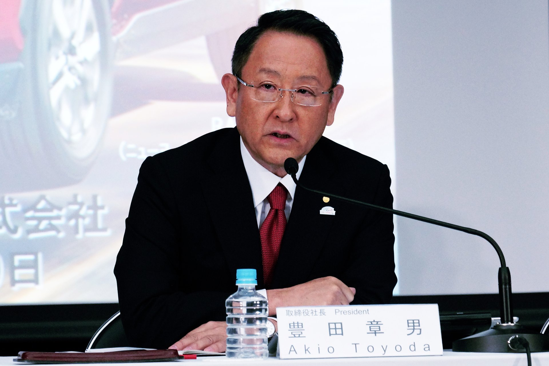 Akio Toyoda is shown seated at a table during a press conference.