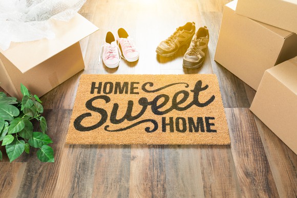 Home Sweet Home Welcome Mat, Moving Boxes, Shoes and a Plant on Hard Wood Floors.