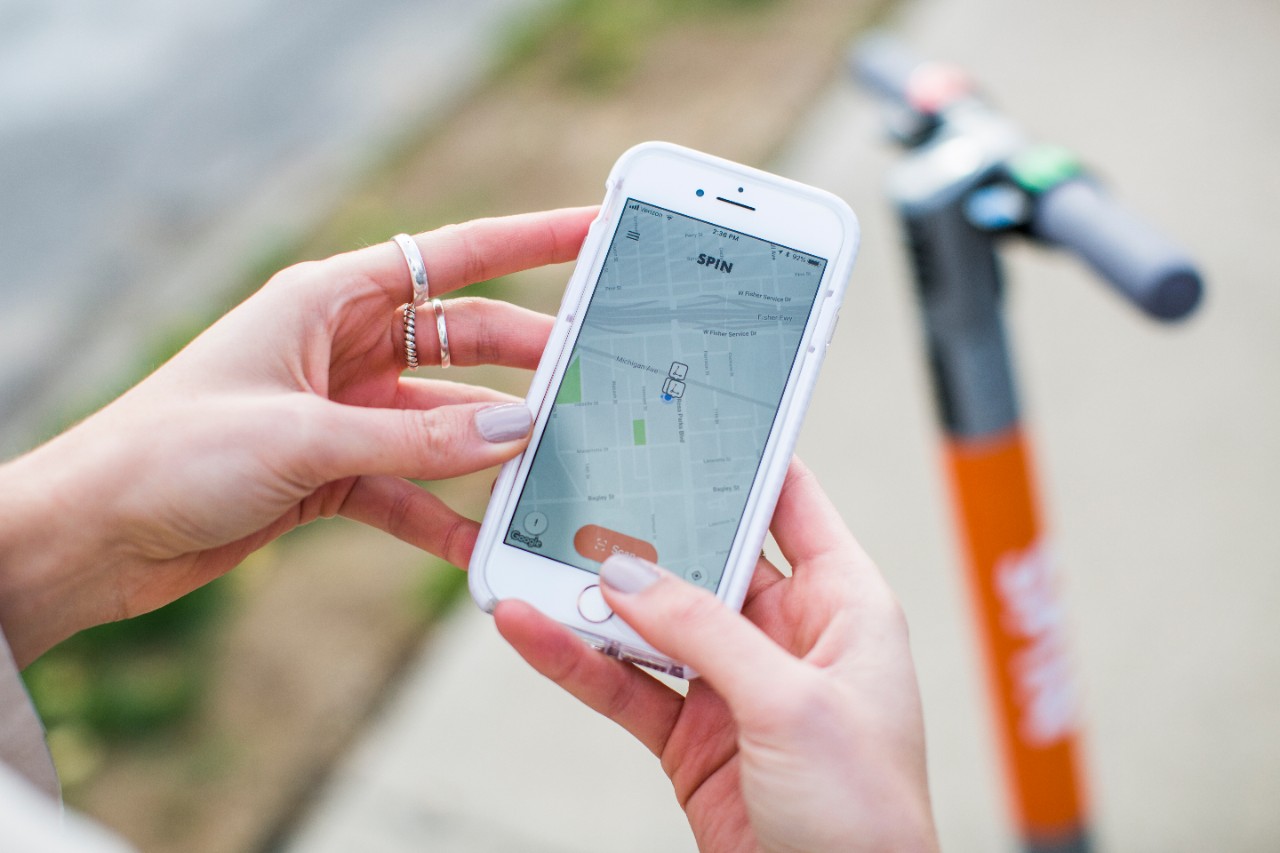 A close-up of a woman's hands holding an iPhone running Spin's app. A Spin scooter is visible in the background.