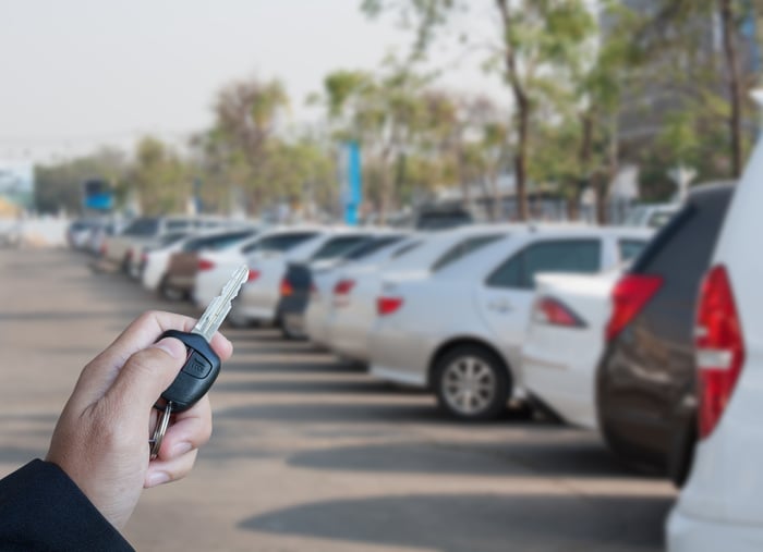 A person's hand holding a car key, with an auto dealer's lot in the background