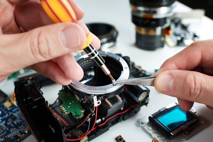 A technician is putting together a camera from parts spread across a white table.