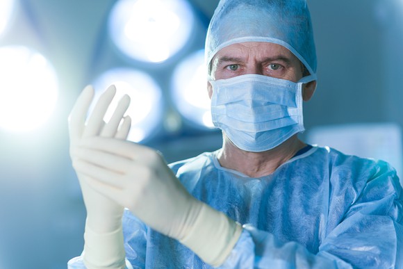 A male doctor with gloved hands and mask over his mouth, as if preparing for surgery