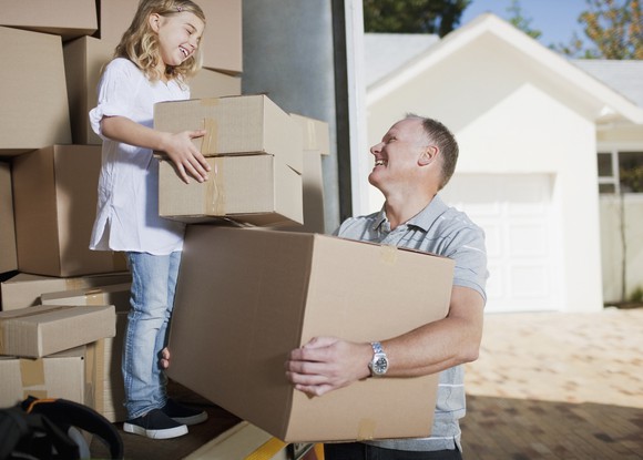 Middle-aged man and young girl carrying boxes from the back of a moving truck with a house in the background.