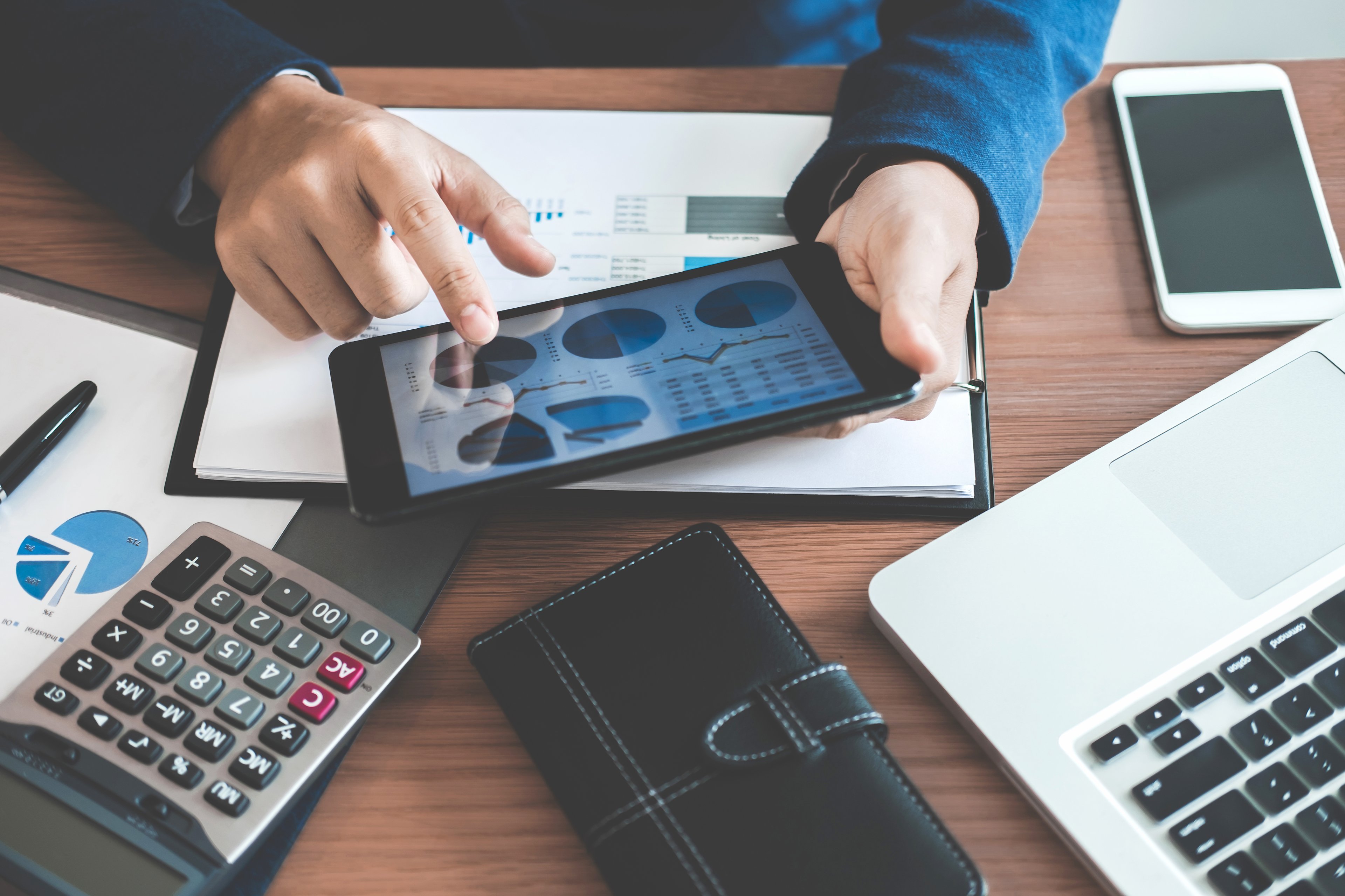 Sitting at his desk, a businessman holds a tablet which displays several financial charts.
