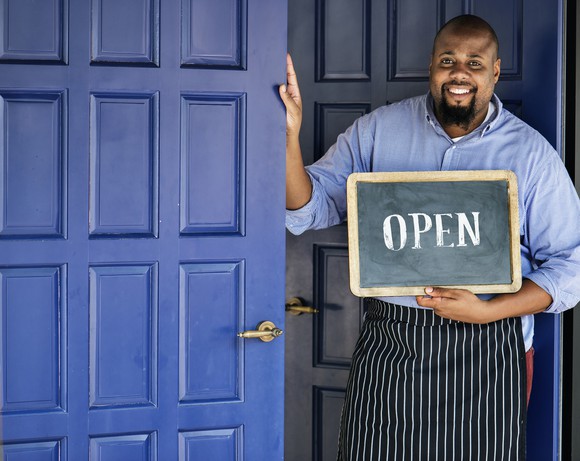 Man standing beside a blue door with a sign saying open.