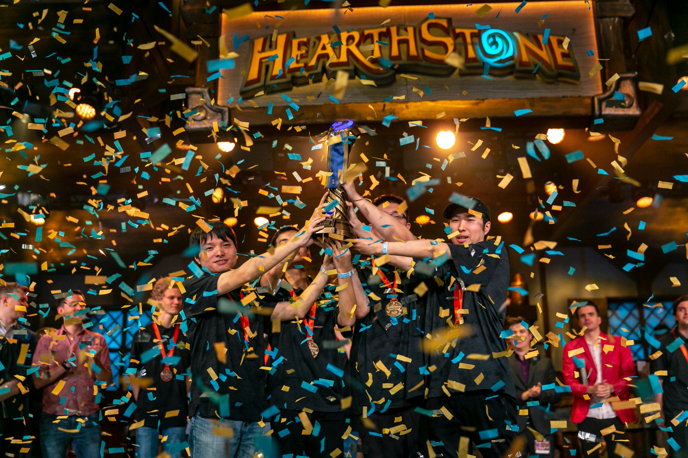 Professional Hearthstone players holding up a trophy with confetti falling as they celebrate a victory at an esport event.