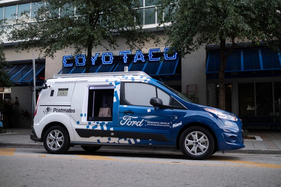 A Ford Transit Connect, a small commercial van, with blue and white Ford graphics and visible self-driving hardware, is parked in front of a restaurant in Miami.