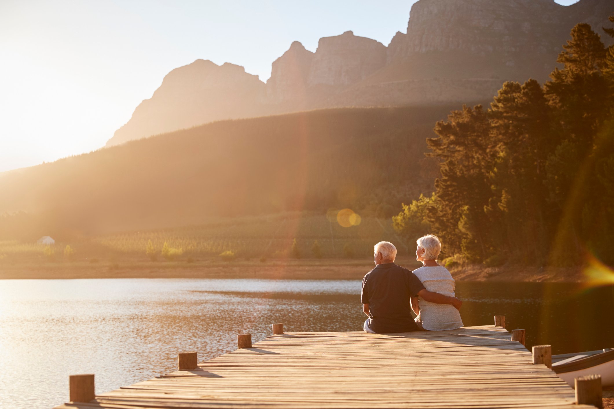 A retired couple sit at the end of a dock on a lake at sunset.