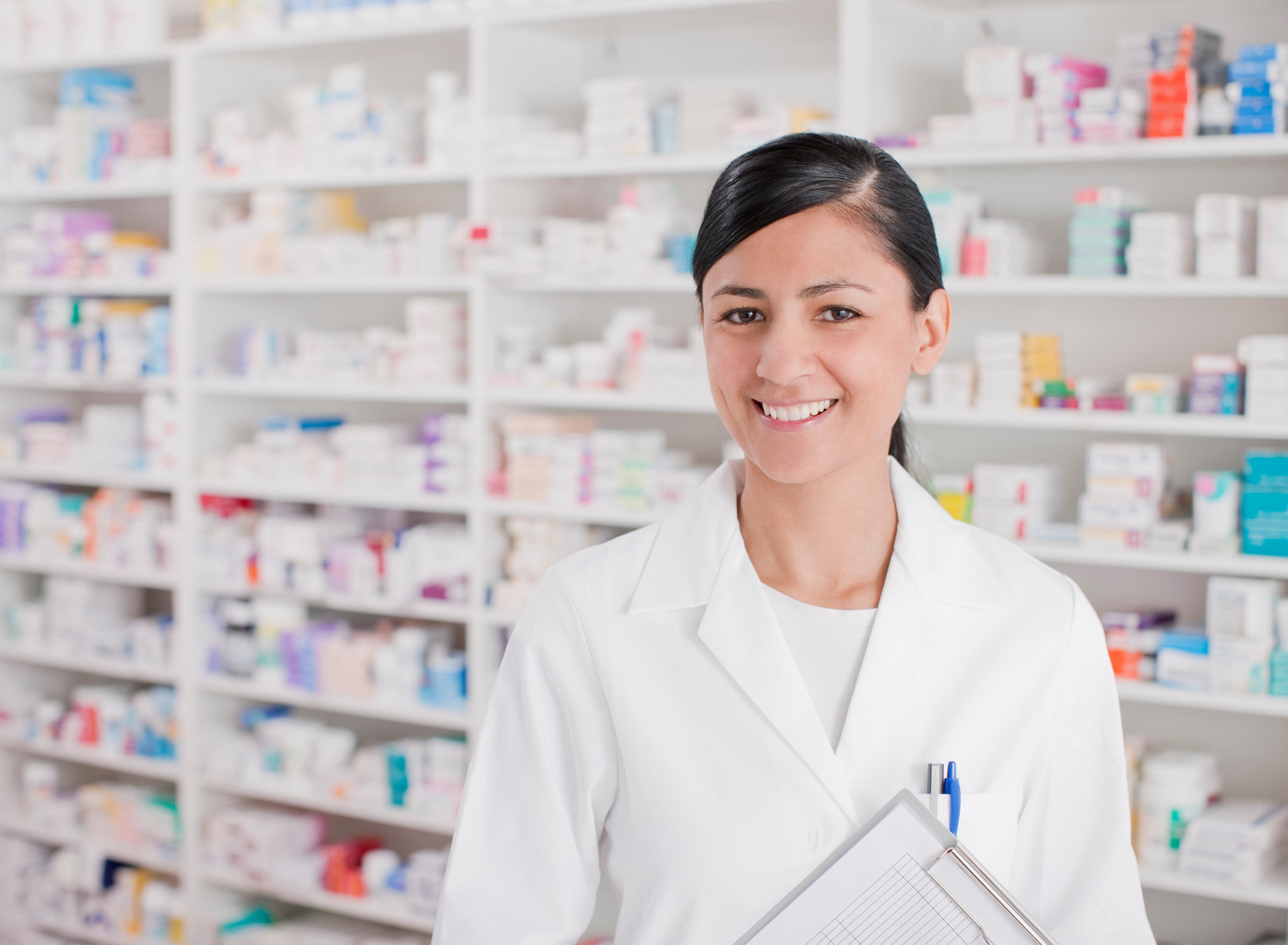 Woman in white lab coat holding clipboard in front of shelves of prescription drug bottles and containers.