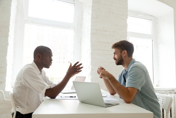 Two men talking at a table with a laptop between them.