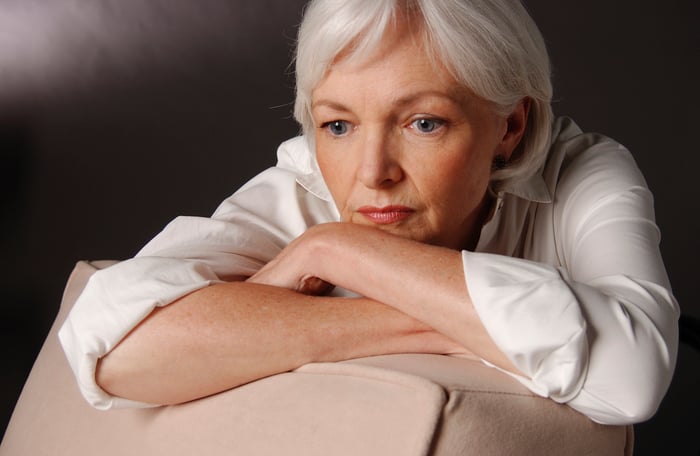 An elderly woman worried, arms crossed and resting on the back of a chair, her head resting on her forearms. 