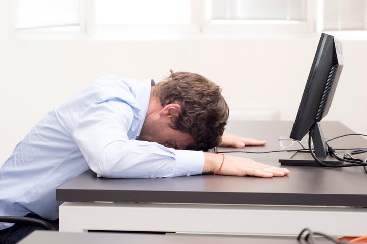 Man with his face down on desk in front of a computer screen.