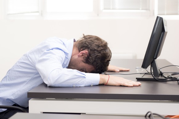 Man with his face down on desk in front of a computer screen.