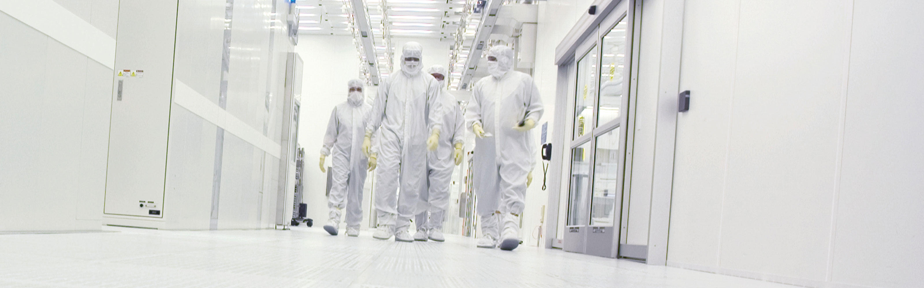 Employees in white full body protective gear walk through the hallways of a research plant. 