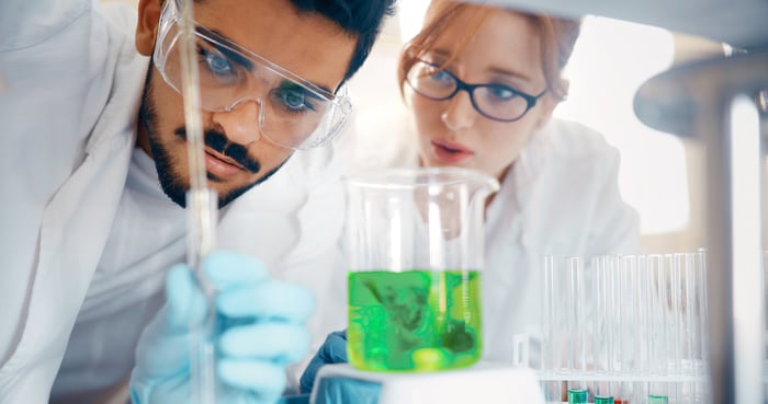 Male and female scientists crouched down looking at dropper, test tube, and beaker containing green fluid.