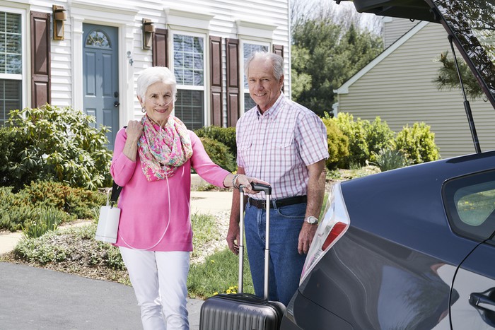 Woman wearing a shoulder-slung Inogen portable oxygen producer while holding a rolling carry-on suitcase and standing next to a senior man and his car.
