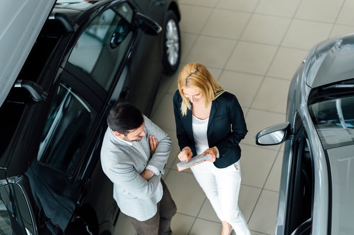 A woman and a man holding papers between two cars at a dealership.