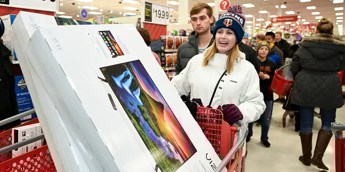 A woman in winter coat and hat pushes a shopping cart with a huge TV in it in Target. 