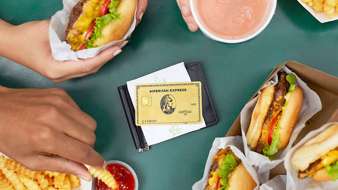 American Express gold card sitting on top of a wallet in the middle of a table with cheeseburgers and fries encircled around it.