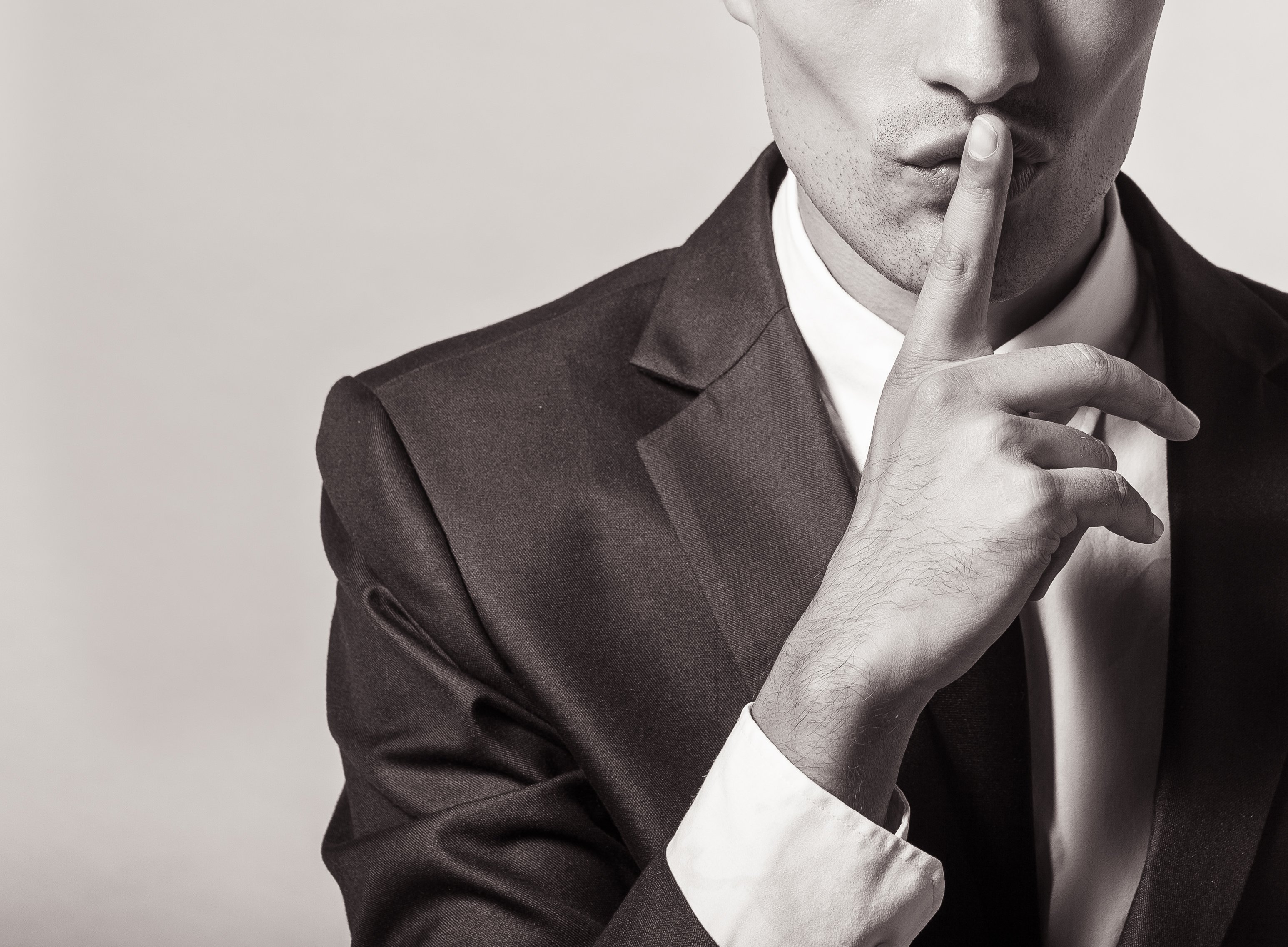 Part of a man in a suit is shown -- we see him holding a finger against his lips, signifying a secret.