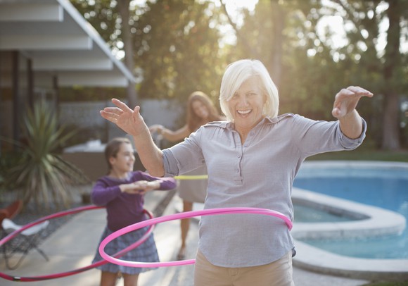 A senior woman using a hula hoop with children playing in the background.
