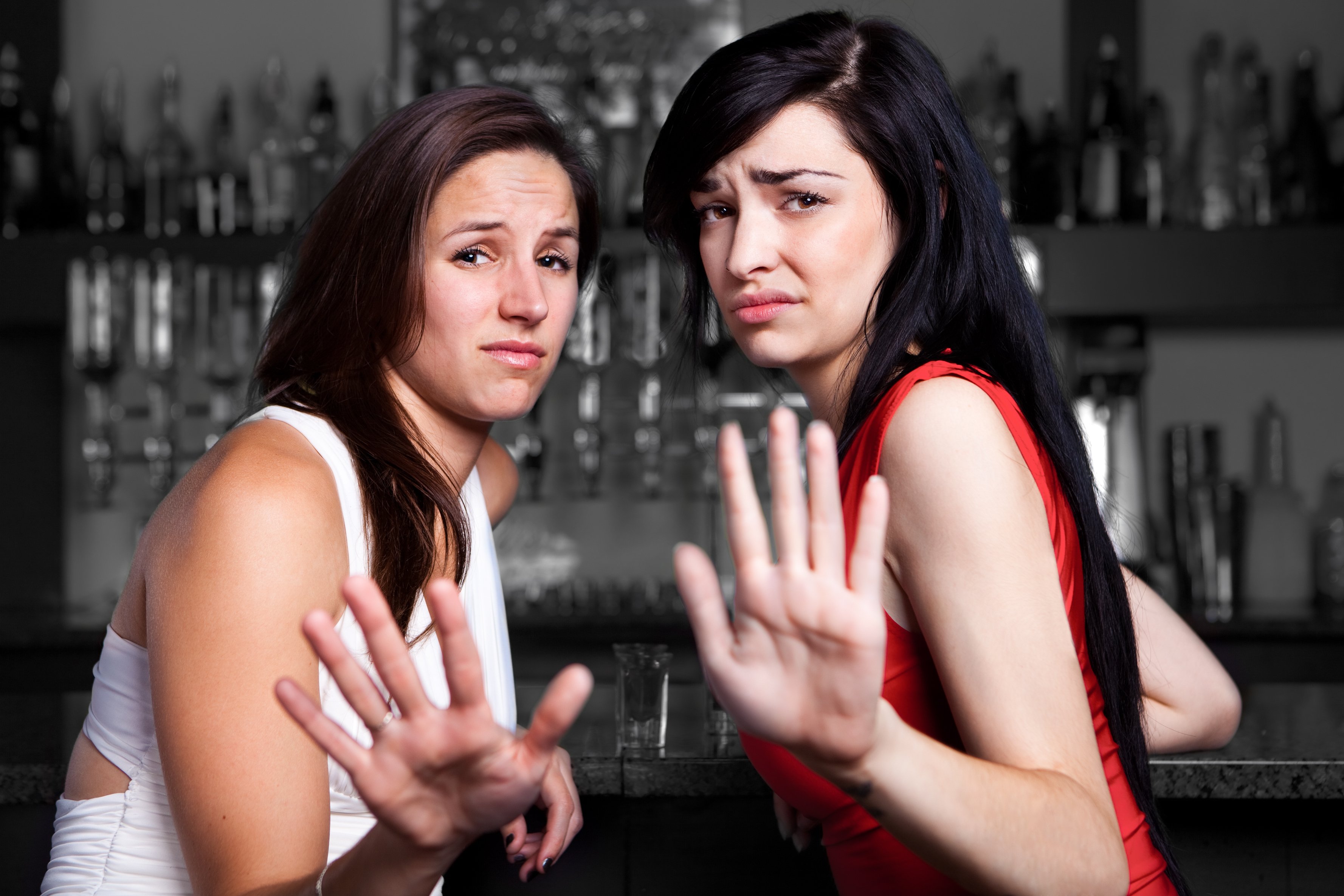 Two young women sit at a bar and hold up their hands, apparently saying no thanks.
