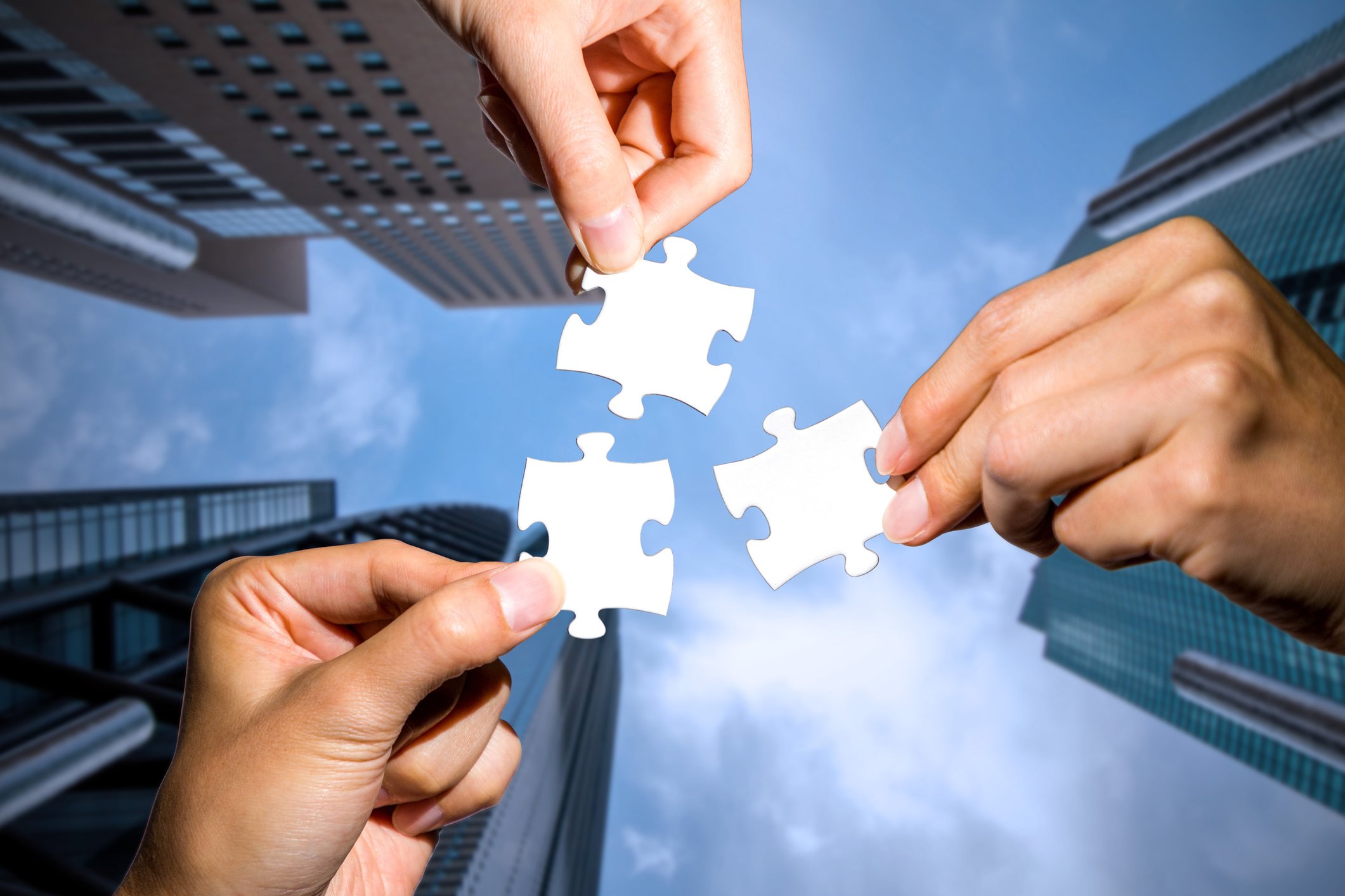 Three hands each holding a white jigsaw puzzle piece against the backdrop of a blue sky and three skyscrapers.