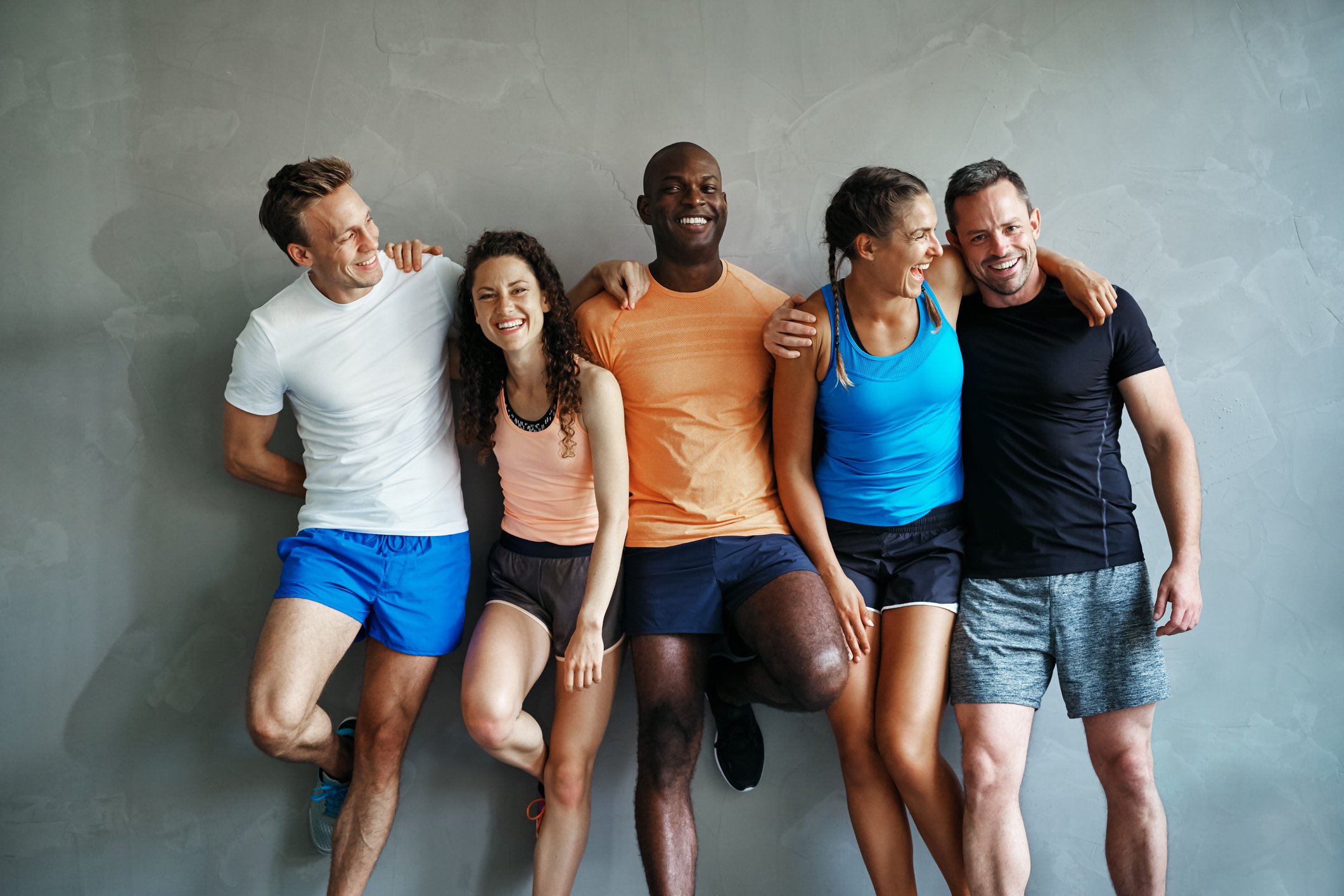 Smiling men and women wearing activewear are standing against a grey wall.