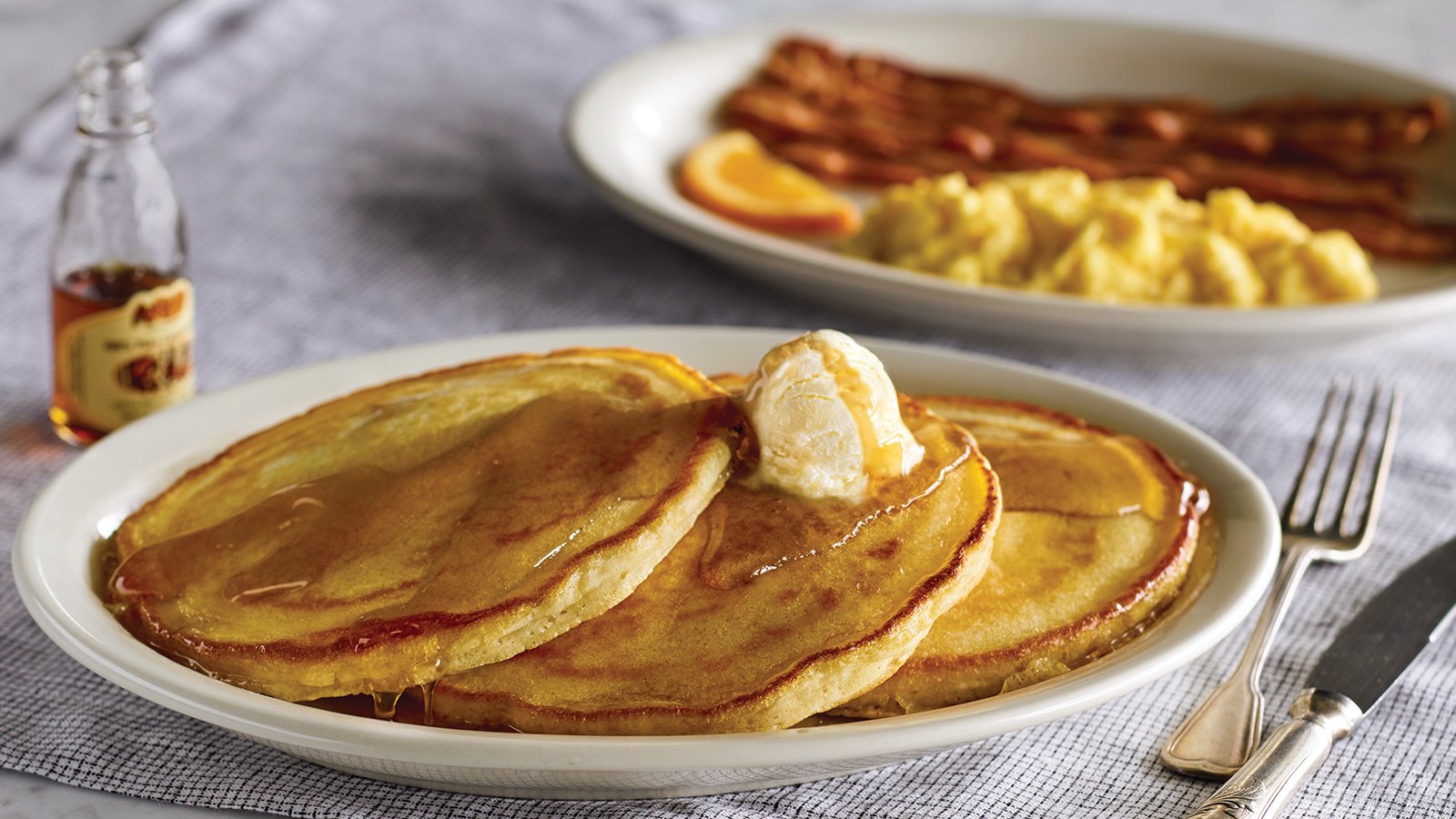 On a tabletop are two plates, One has eggs and bacon, and the other has pancakes with butter and syrup.
