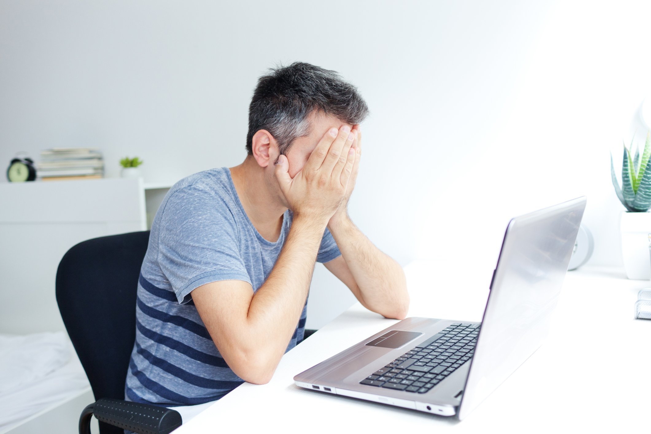 Man covering his face while sitting at a desk with an open laptop